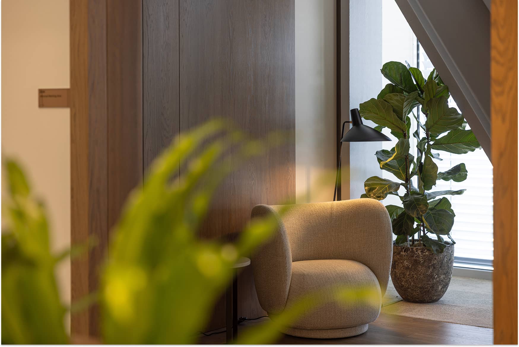 Cozy corner with a beige armchair, potted ficus plant, and floor lamp by a window, framed by wooden panels and blurred greenery in the foreground.