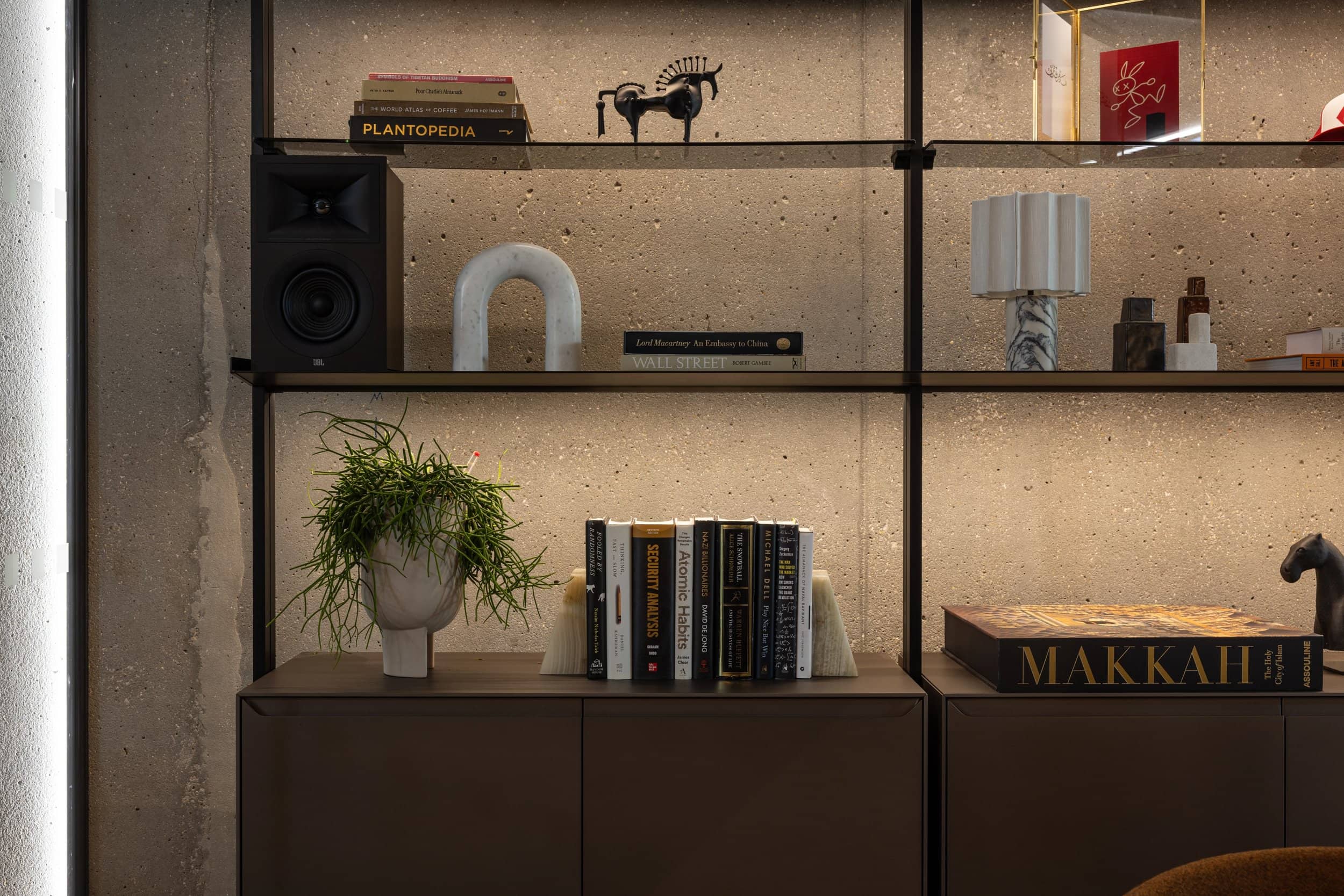 A modern shelf with books, a speaker, decorative items, a potted plant, and a lamp against a textured wall.
