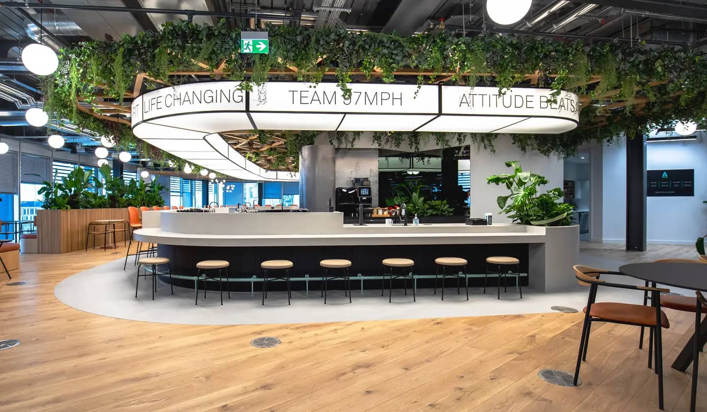 Modern office break area with a circular bar, stools, hanging plants, and motivational signs. Bright lighting and wooden flooring.