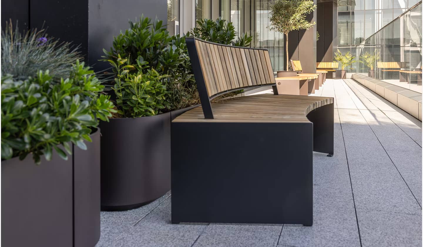 An outdoor terrace, with outdoor planters, and a sleek bench. An olive tree can be seen in the background